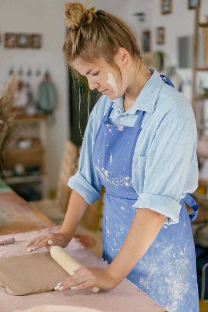 A woman in a blue apron concentrates on crafting pottery in a workshop, showcasing handmade skills.